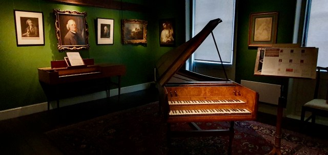 "Harpsicord, like a grand piano but smaller, the top lid open and on the green walls many paintings."