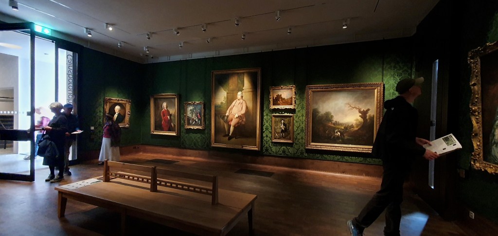 "Green silk woven gallery walls and on these a various set of large and smaller paintings by Gainsbrorough. Quite dark and wooden floor."