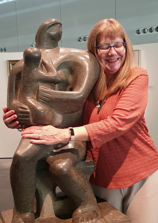 'Bronzed-coloured modern art sculpture of mother and child, mother's head turned towards mine as I embrace them both, my head resting on mother's shoulder.'